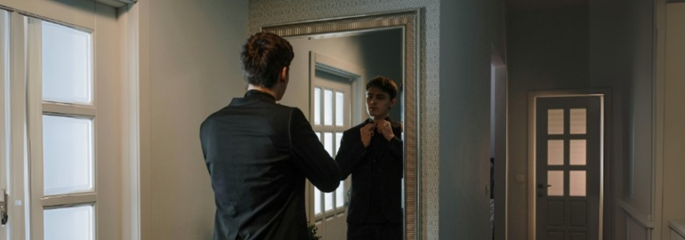 A fresh graduate getting ready for a job interview in front of a mirror wearing a tie.