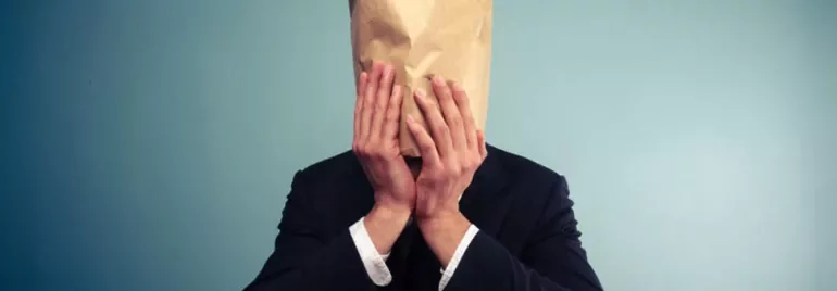 Person in a suit with a paper bag over their head against a blue background.