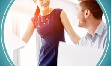 Two colleagues smiling and discussing work at a desk in a bright office.