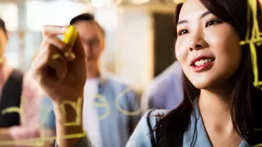 Woman writing on transparent board with yellow marker, people in background.