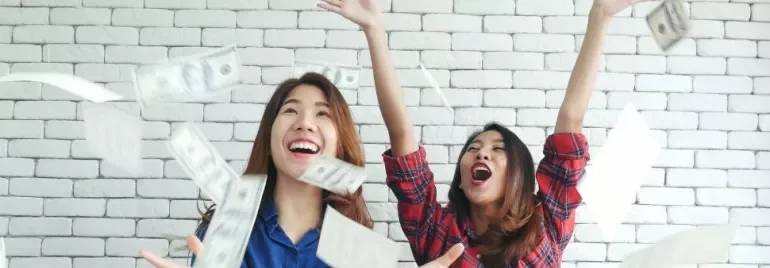 Two women happily throwing money in the air against a white brick wall.