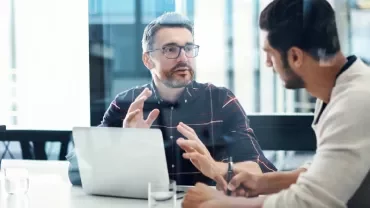 Two men discussing at a table with a laptop in an office setting.