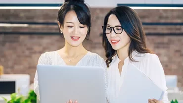 Two women smiling and looking at a laptop in an office environment.