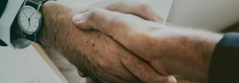 Close-up of a handshake over a wooden desk with documents and a laptop in the background.