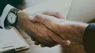 Close-up of a handshake over a wooden desk with documents and a laptop in the background.