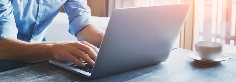 Person typing on a laptop at a wooden table with a cup of coffee nearby.
