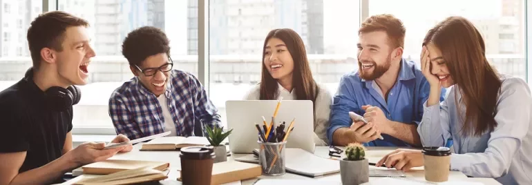 Diverse group of students laughing and studying together with laptops and coffee in a modern setting.