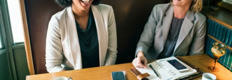 Two women smiling and chatting at a cafe table with papers and coffee.