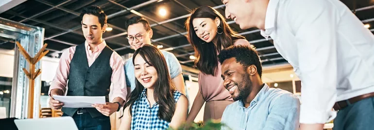 A group of six colleagues smiling around a laptop in an office.