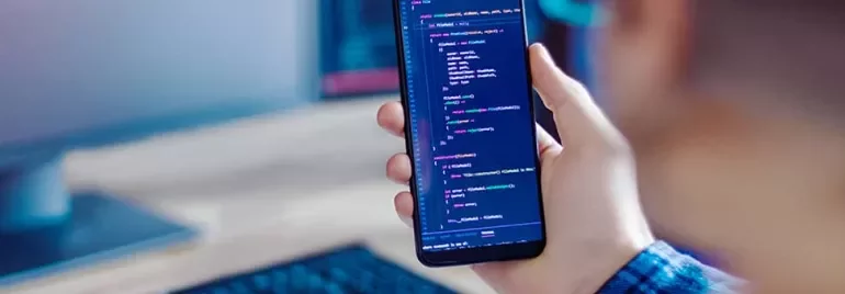 Person holding a smartphone displaying colorful code, with a blurred computer screen in the background.