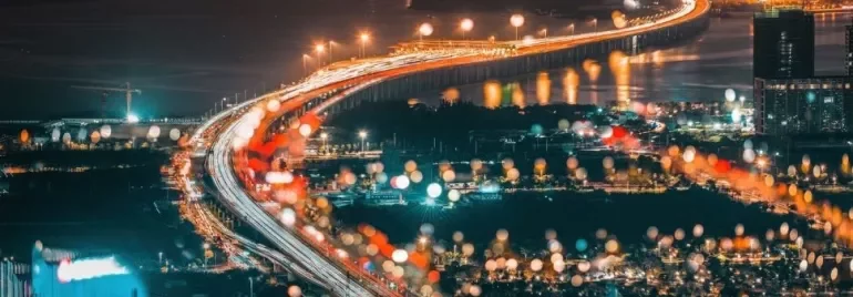 Aerial view of a city at night with a brightly lit bridge and blurred lights in the foreground.