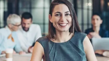 Smiling woman in an office setting with colleagues working in the background.