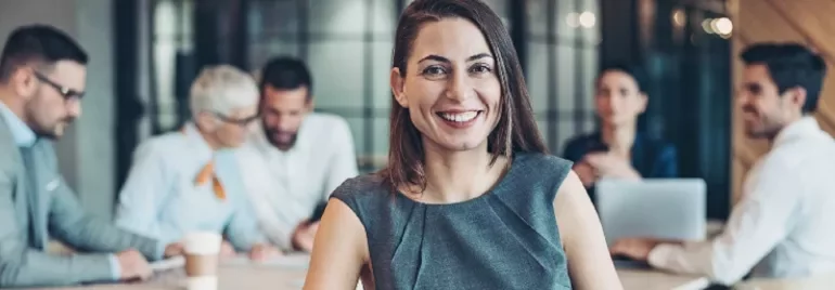 Smiling woman in an office setting with colleagues working in the background.