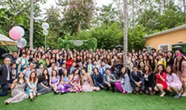Large group of people posing on a green lawn with trees and balloons in the background.