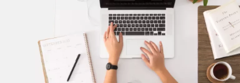Overhead view of person working on a laptop with a planner, plant, and cup of coffee on the desk.
