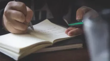 Person writing notes in a notebook while holding a smartphone.