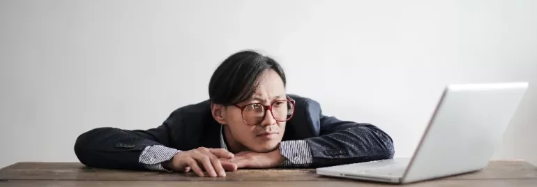 Man in suit and glasses staring thoughtfully at an open laptop on a wooden table.