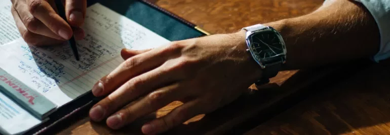 Person writing notes with a black pen at a wooden desk.