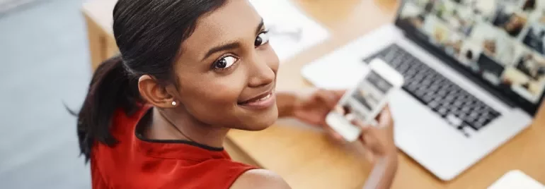 Woman in red top smiling, holding a smartphone while looking at a laptop on a wooden desk.