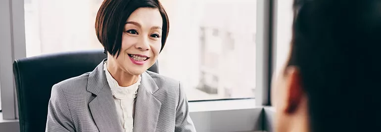 Woman in a gray suit talking to another person in an office setting.