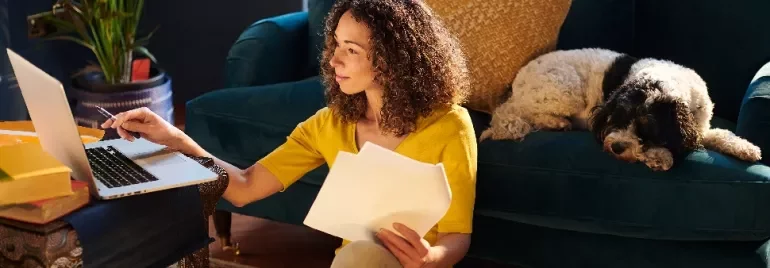 Woman working with a laptop and papers, dog resting on couch in sunlit room.