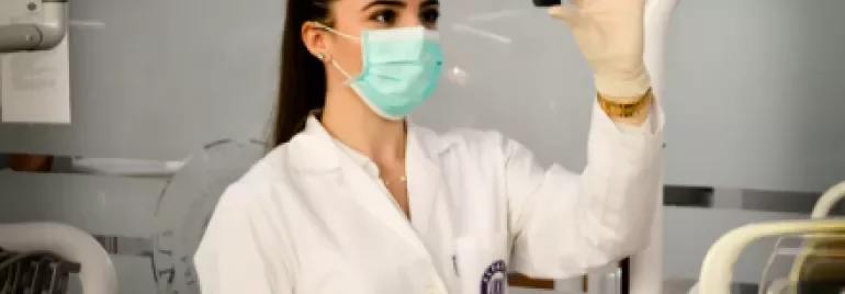 Scientist in lab coat and mask examining a small object in a laboratory.