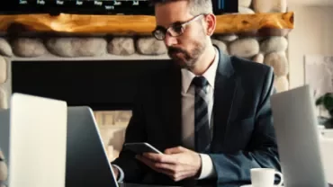 Man in suit working on laptop and smartphone at a table with a coffee cup.