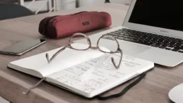 Open notebook with glasses on a desk near a laptop, phone, and pencil case.