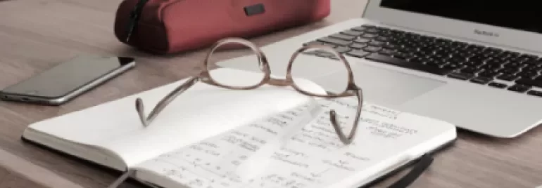 Open notebook with glasses on a desk near a laptop, phone, and pencil case.