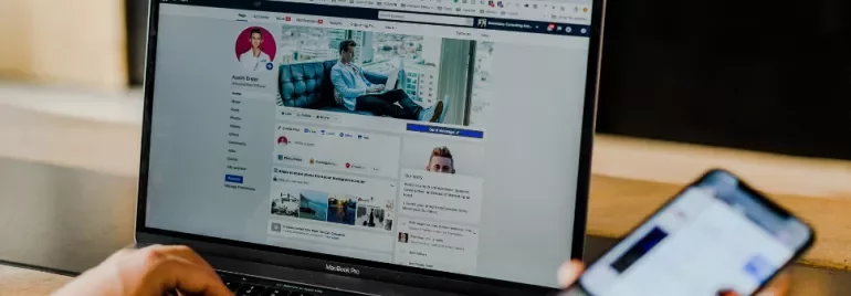 Person using a laptop, browsing a social media profile, while holding a smartphone.