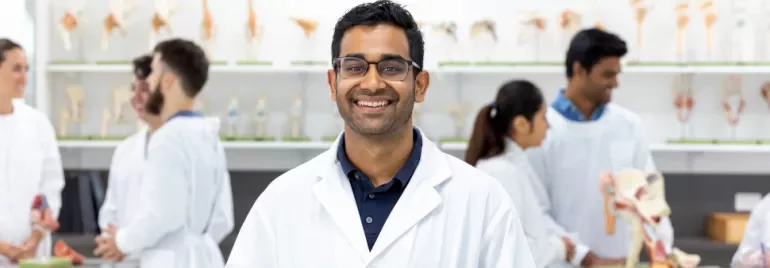 A smiling lab technician in a white coat stands in a science classroom with colleagues working in the background.