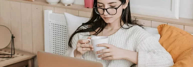 Woman in glasses reading on a laptop and holding a mug