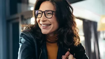Woman in glasses smiling while seated indoors using a laptop.
