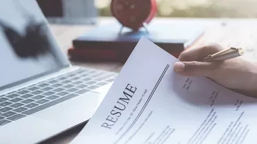 Person holding a resume next to an open laptop on a desk.