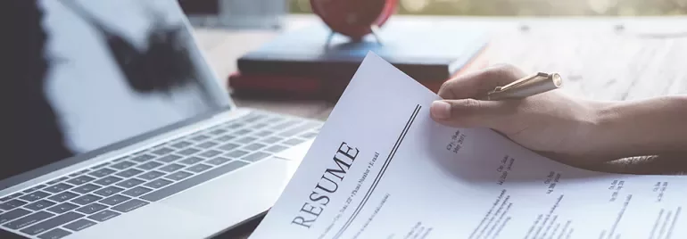 Person holding a resume next to an open laptop on a desk.