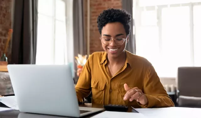 Person smiling while working on a laptop and calculator in a bright office.