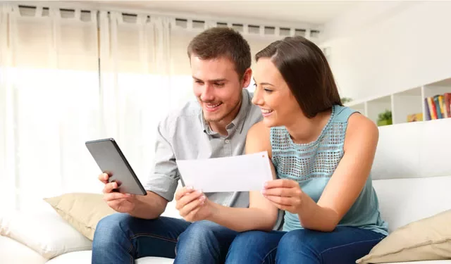 Couple sitting on couch, smiling at tablet and holding paper.