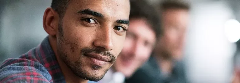 Man in a plaid shirt smiling with colleagues in a blurred background.