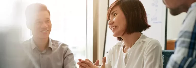 Three people having a discussion around a table with a whiteboard in the background.