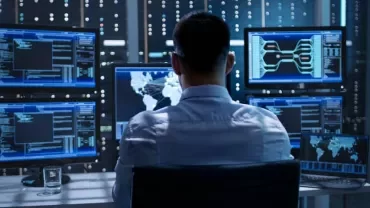 Person working in a data center, using multiple monitors displaying code and world maps.