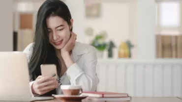 Woman smiling at phone in a cozy cafe with a laptop and coffee.