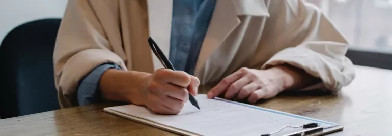 Person signing a document on a clipboard at a wooden table.