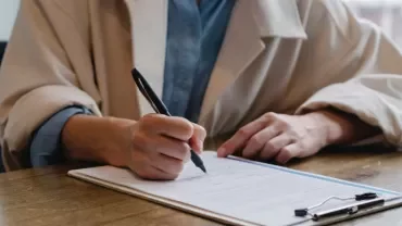 Person signing a document on a clipboard at a wooden table.