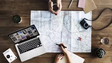 Map, laptop, camera, and notebooks on a wooden table with hands planning a trip.