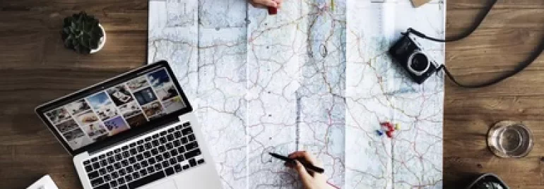 Map, laptop, camera, and notebooks on a wooden table with hands planning a trip.