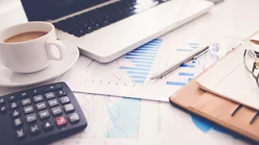 Laptop on desk with charts, calculator, coffee cup, and notebook.