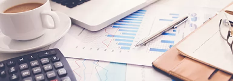 Laptop on desk with charts, calculator, coffee cup, and notebook.