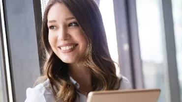 Smiling woman holding a tablet, gazing out of a large window.